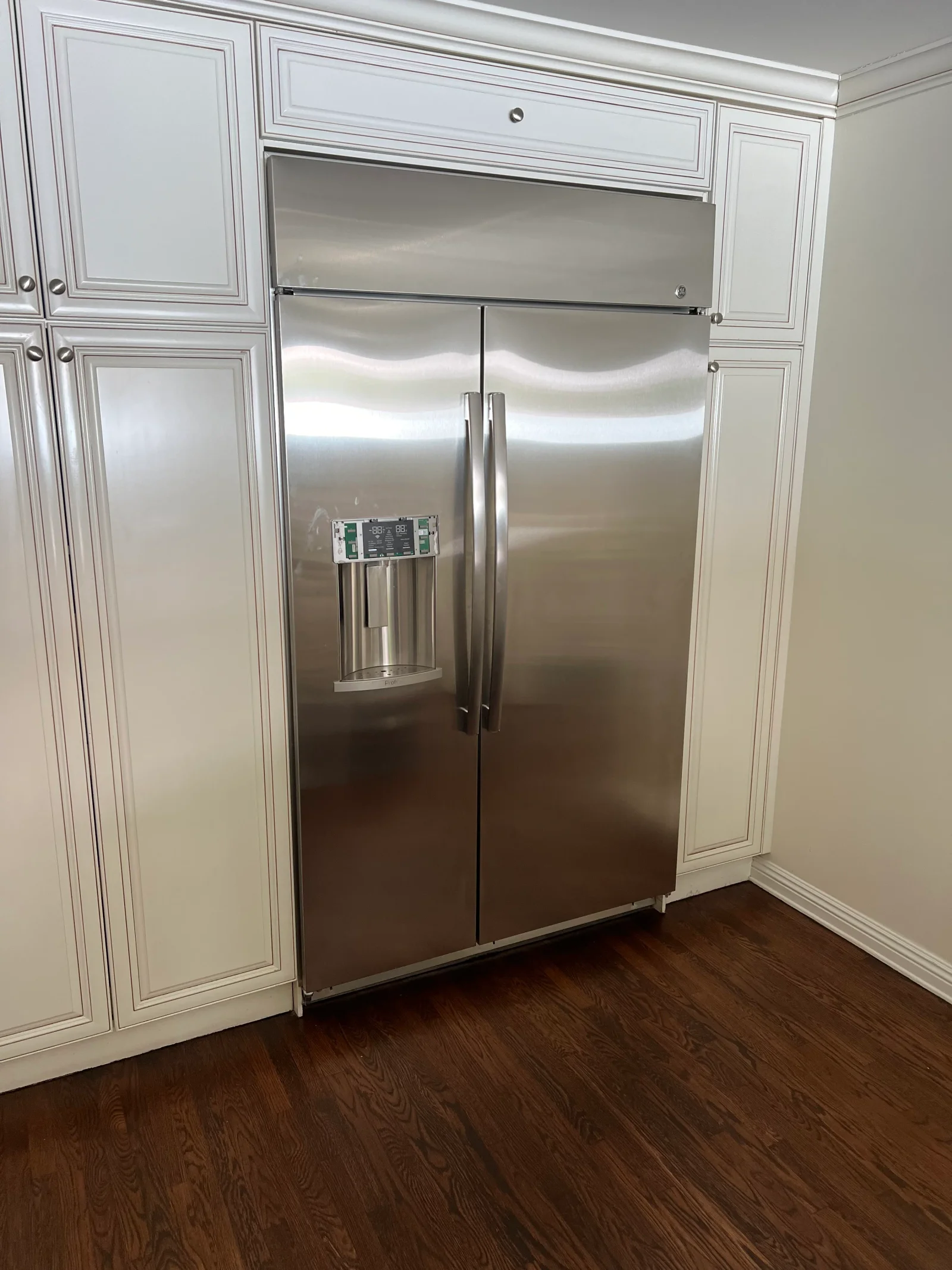 Stainless steel built-in refrigerator with ice dispenser installed between white painted kitchen cabinets