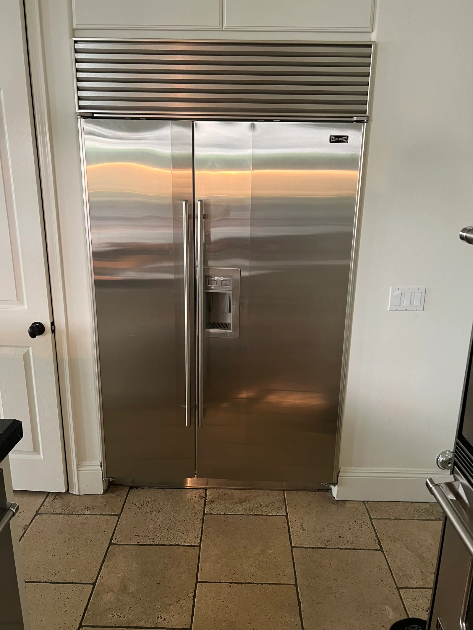 Stainless steel built-in refrigerator with dual doors, chrome handles, and motorized louver vent above