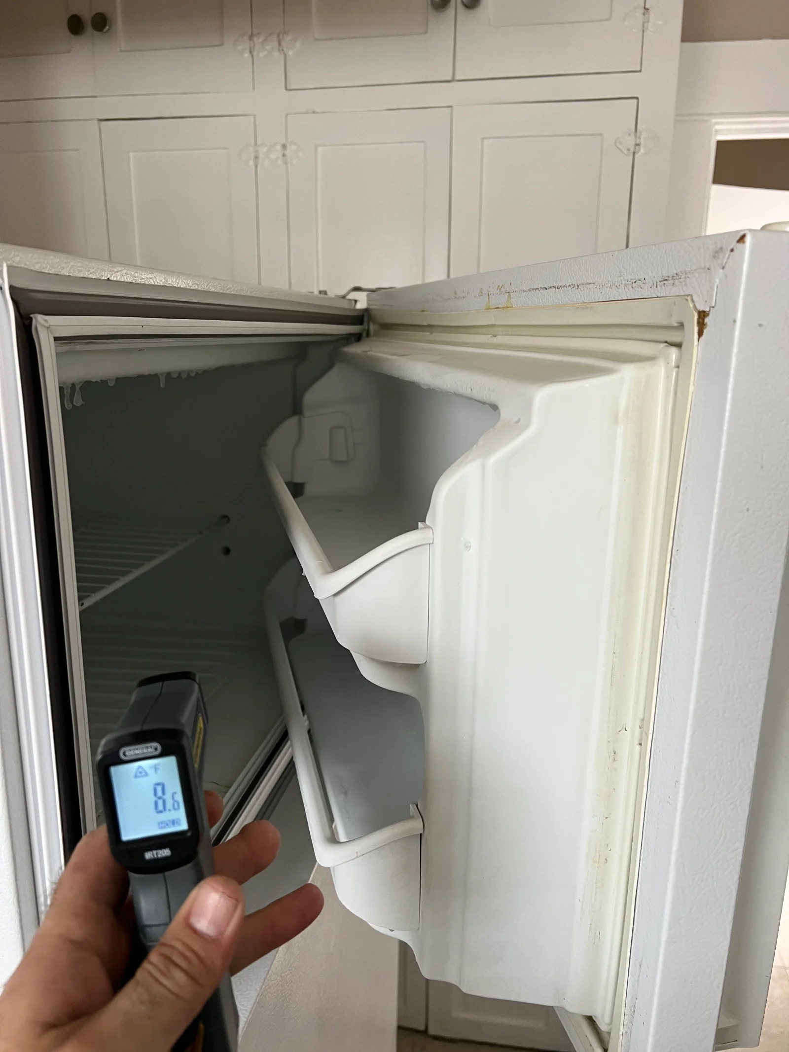 Open refrigerator interior with diagnostic thermometer measuring temperature, white shelving and cooling components visible